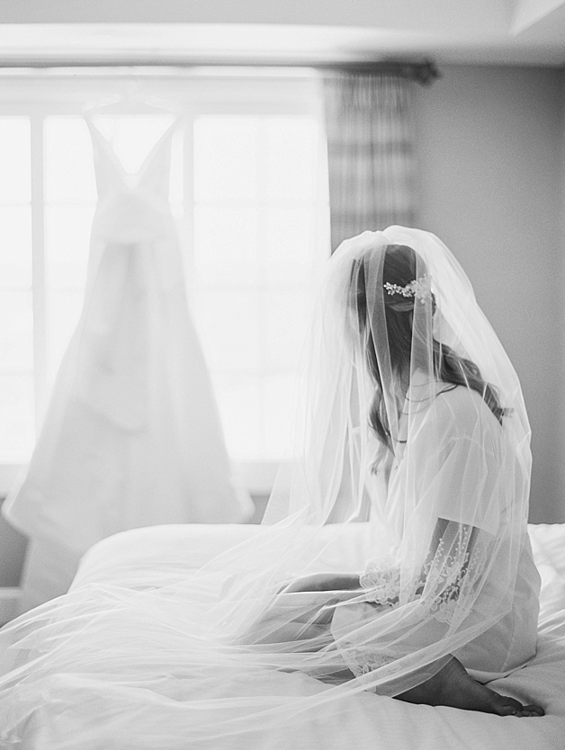 Bride gazing at her wedding gown in veil on bed
